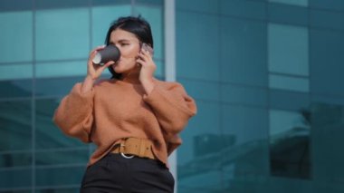 Mutlu İspanyol kadın dışarıda durmuş kağıt bardaktan kahve içiyor arkadaşça bir gülümsemeye karşılık veriyor cep telefonuyla arkadaşlarıyla uzaktan iletişim kuruyor.