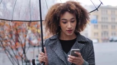 Sonbaharda açık havada yağmurda şeffaf şemsiyesi olan Afro-Amerikan kıvırcık kadın telefonuyla iyi haberlerle kazanıyor. İnternet üzerinden bahis yaparak zafer kazanmanın mobil mutluluğunu sunuyor.