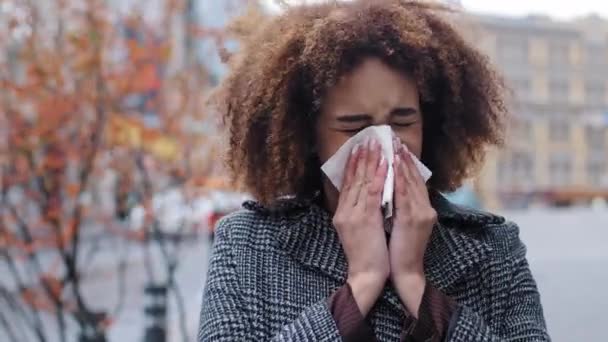 Doente afro-americana doente com cabelo encaracolado soprando corrimento nasal em papel tissue ao ar livre alergias frias vírus respiratórios menina sofre de rinite contagiosa sintoma outono na cidade gripe pandêmica — Vídeo de Stock