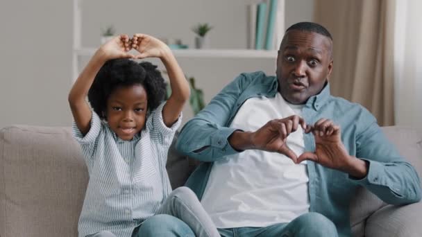 Feliz divertido afroamericano familia madura padre e hija pequeña sentado en el sofá en la habitación sonriendo hacer que la forma del corazón con los dedos muestran gesto amor símbolo cuidado signo padre-niño relación concepto — Vídeos de Stock