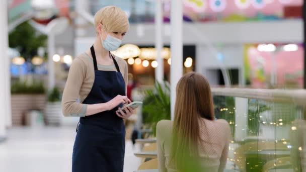 Kvinnlig servitris anställd arbetstagare i cafeteria restaurang i förkläde och medicinsk mask serverar samtal med kvinnliga kund kund flicka sitter vid bordet skriver ordning i mobiltelefon råd om mat lunch skålen — Stockvideo