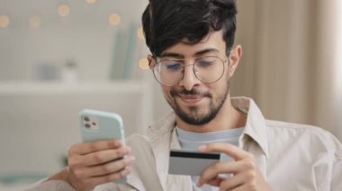 Yakın plan sakallı, kredi kartı ve telefon ile gözlüklü bir Arap işadamı online alışveriş yapıyor. Banka numarası çevrimiçi işlem için mobil finansal uygulama rezervasyon siparişi kullanıyor.