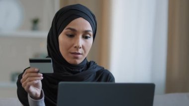 Tesettürlü çekici bir kız elinde kredi kartı ödemesi internet siparişi, bankacılık başvurusunu dizüstü bilgisayarda gülümseyen müslüman kadın alışverişi başarılı bir şekilde seviniyor güvenli para transferi modern teknoloji konsepti