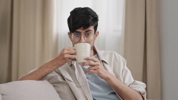Arabský vousatý tisíciletý muž latinsky chlap v brýlích sedící doma a vychutnávající si lahodné horké ovoce bylinný čaj káva nápoj kakao pití cappuccino nápoj říká ano pozitivně kývnout hlavou útulnost koncept — Stock video