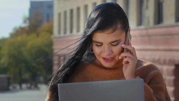 Potret perempuan muda hispanik gadis multitasking duduk di luar ruangan melihat layar laptop berbicara di ponsel menjawab panggilan ramah memesan di toko online belajar jarak jauh menggunakan aplikasi — Stok Video