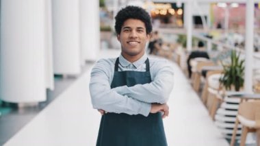 Genç Afro-Amerikalı adam, restoran çalışanı, kafeterya çalışanı, kameraya bakıyor. Önlük giymiş, iş yerinde gülümseyen bir garson pozu veriyor. Küçük işletme