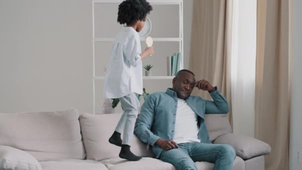 Mature african american exhausted tired after work father sitting on sofa little cheerful noisy daughter jumping on couch interferes disturbs dad from resting man upset by behavior child kid girl play — Stock Video