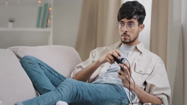 Arabiske tusindårige fyr skæg indisk mand bærer briller liggende sofa spille konsol nyde online videospil konkurrence vindende gør ja hånd gestus have det sjovt brug controller hobby videospil koncept – Stock-video