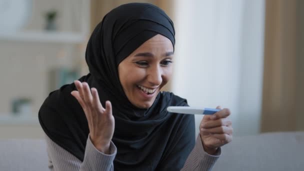Портрет радісної здивованої дівчини в хіджабі, що тримає тест на вагітність, сидить у вітальні з позитивним результатом красива ісламська молода жінка відчуває себе щасливою після успішного лікування фертильності концепції IVF — стокове відео