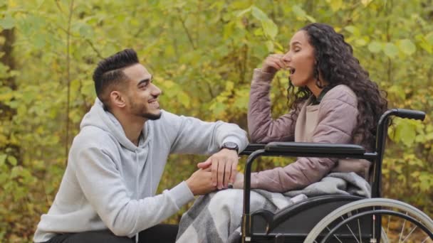 Fiatal spanyol férfi guggoló beszél lány tolószékben kéz a kézben pár szerelmes pihentető természetben srác látogatás beteg barátnő a kórházi parkban rehabilitáció alatt gondoskodó fogyatékkal élő beteg — Stock videók