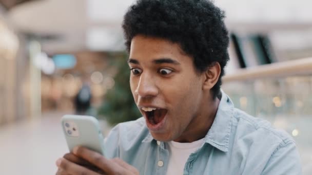 Africký Američan překvapený obdrženou zprávou vyhrává on-line sázky při pohledu na mobilní telefon otevírá ústa v překvapení potěšený šok muž cítí štěstí z nečekané novinky úžasná nabídka příležitost — Stock video