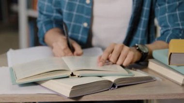 Yakın çekim, tanınmayan genç erkek öğrenci kütüphanede oturmuş ödevlerini yapıyor sınav sınav notları hazırlıyor ders kitabından notlar yazıyor kâğıda yazılmış bilgileri dikkatlice okuyor.