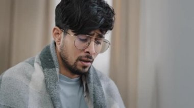 Yakın plan yorgun Arap sakallı adam mevsimsel alerji tozu virüsü solunum hastalığına yakalanmış gözlüklü adam battaniyeye sarılmış, evinde oturmuş kağıt peçeteye sümük sümküren adam.