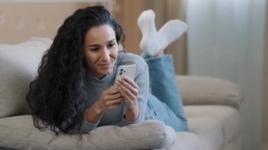 Çekici 25 'li yaşlarda kız kanepede uzanmış telefonda dinleniyor internette sohbet ediyor gülümseyen genç bir kadın hafta sonunun keyfini çıkarıyor internetten neşeli esmer, kaygısız bir şekilde sosyal medyada geziniyor.