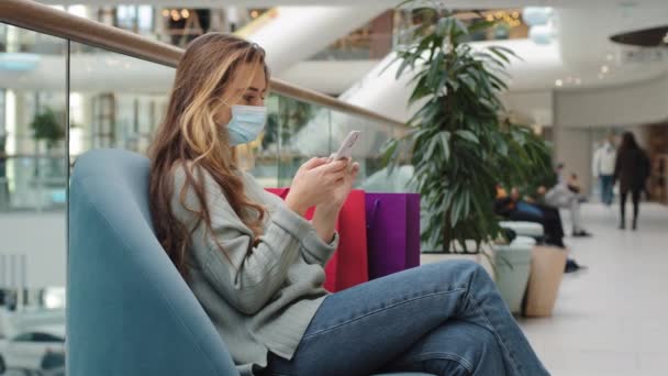 Кавказская женщина покупатель носить медицинскую маску расслаблены с покупками сумки женщина больная покупатель сидит в помещении на диване в магазине глядя на мобильный телефон покупая онлайн приложение выбирая продукт чата бесплатный Wi-Fi — стоковое видео