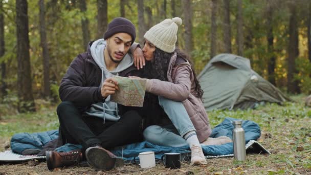 Happy young indian couple hikers sit in nature near tent look at paper map check route directions using guidebook two travelers in love on romantic holiday trip tourist travel concept with backpack — Vídeo de Stock