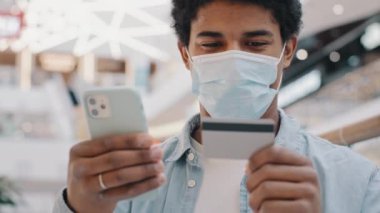Tıbbi maskeli Afrikalı Amerikalı müşteri telefon mobil uygulaması ile bankacılık kredi kartı verilerini girerek mal satın alıyor internet hizmetleri ödemeleri kolay para transferleri