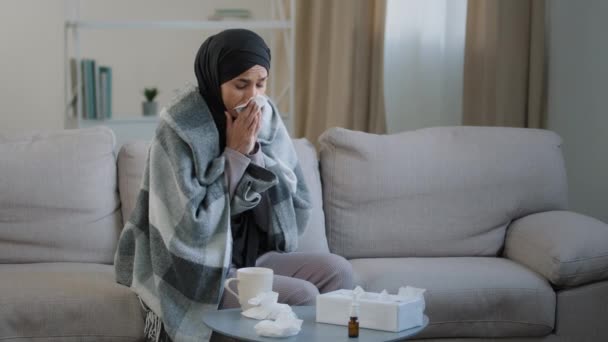 Beteg fiatal nő fekete hidzsáb csomagolva magát takaró ül a kanapén a nappaliban beteg iszlám lány tüsszent törli orrát szalvéta ivás forró tea érzés fejfájás hőmérséklet allergia tünetei — Stock videók