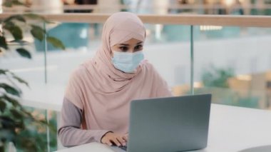 Tıbbi maske takan Arap kadın laptopa bakıyor. İyi haberler okuyor. Zaferi kutluyor.