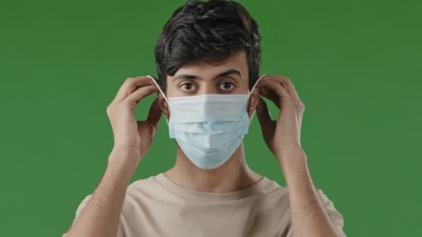 Ciddi bir Arap genç adam, tıbbi maskesini takıp yeşil arka planda duruyor ve Covid19 Coronavirus Salgın Önleme Konsepti 'nin kişisel koruyucu ekipmanlarına bakıyor. — Stok video