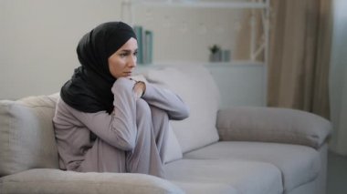 Siyah tesettürlü üzgün genç bir kadın oturma odasında kanepede oturup psikolojik sorunlardan muzdarip evde hayal kırıklığına uğramış Müslüman bir kız stresli, olumsuz düşüncelere dalmış.