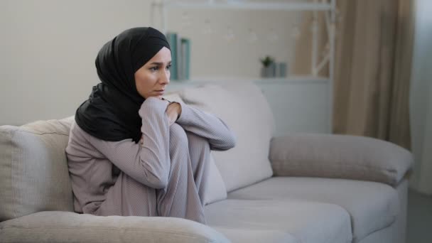 Mujer joven triste ansiosa en hiyab negro sentado en el sofá en la sala de estar que sufre de problemas psicológicos problemas en el hogar frustrado chica musulmana sensación de estrés deprimido perdido en pensamientos negativos — Vídeos de Stock
