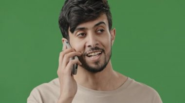 Mutlu gülümseyen genç Arap stüdyoda yeşil arka planda konuşan akıllı telefon kullanan, pozitif adam cep telefonu modern teknoloji kavramında arkadaşlarıyla hoş sohbetler yapıyor.