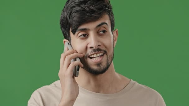 Close up happy smiling young arab guy in studio on green background talking using smartphone surprised positive man chatting with friend pleasant conversation on mobile phone modern technology concept — Stock Video