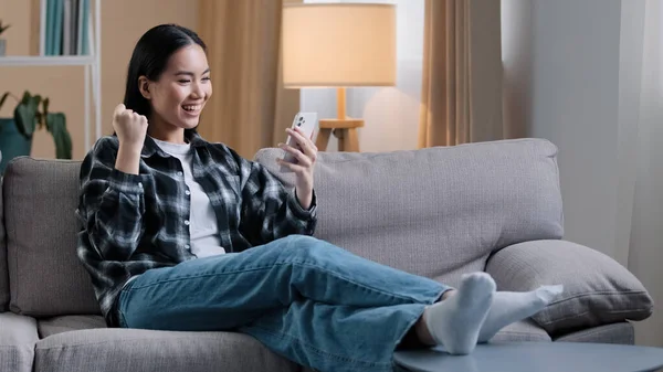 Asiatisk flicka kvinna sitter på soffan avkopplande hemma på soffan spela mobilspel vinner få nytt jobb erbjudande meddelande online betting läser goda nyheter telefon framgång lycka ja seger gest med smartphone — Stockfoto