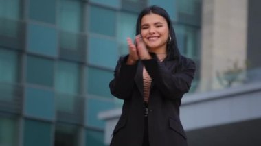 Genç, başarılı, gülümseyen İspanyol iş kadını arka plandaki ofis binasına el çırpıyor. Alkışlara bakıyor.
