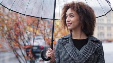 Gülümseyen mutlu Afro-Amerikan 20 'li yaşlarda model, şeffaf şemsiyeli turist kız, şehirde duruyor, sonbahar yağmurunda yağmur yolculuğunun keyfini çıkarıyor, başka yerlere bakıyor, hayal kuruyor.