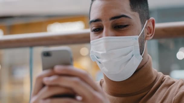 Κοντινό πλάνο νεαρός χαρούμενος Άραβας με ιατρική μάσκα κρατώντας κινητό τηλέφωνο χαμογελώντας σερφάροντας στο διαδίκτυο σχετικά με τη χρονολόγηση site έλεγχο e-mail γράφοντας μήνυμα συνομιλίας με φίλο κάνοντας τάξη στο ηλεκτρονικό κατάστημα χρησιμοποιώντας smartphone — Αρχείο Βίντεο