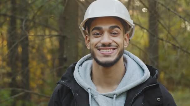 Muž portrét mladý usmívající se hispánec profesionální pracovník v bezpečnostní přilbě pózování venku při pohledu na kameru šťastný sebevědomý muž lesnictví inženýr stojí v dřevě koncepce ochrany přírody zalesňování — Stock video