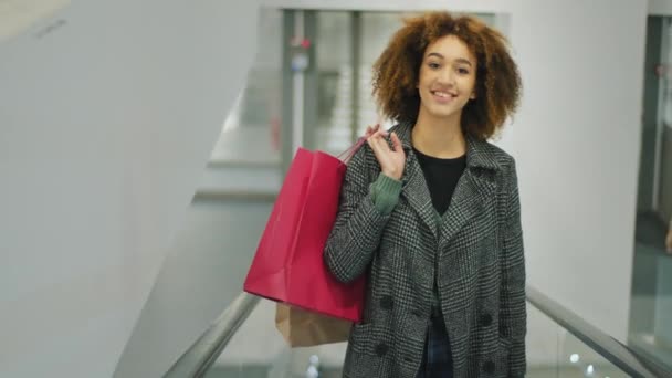 Top vista elegante sorrindo afro-americano menina comprador comprador cliente sorriso feliz descontos mulher com cabelos cacheados em pé na escada rolante em movimento no shopping com compras sacos de compras — Vídeo de Stock