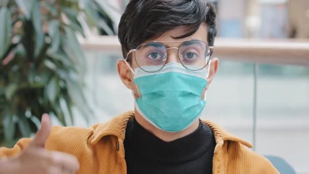 Close-up jovem indiano em máscara médica posando dentro de casa olhando para a câmera mostrando polegares para cima sinal aprovação acordo gesto aprovando símbolo positivo opinião proteção precauções durante a pandemia — Vídeo de Stock