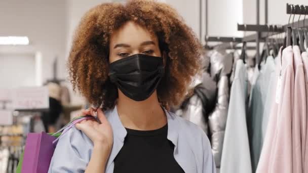 Afro-Amerikalı tüketici kız tüketici alıcı kıvırcık kadın müşteri giyim mağazasından alışveriş çantası satın alma indirimi karantinası ile tıbbi koruyucu maske takıyor. — Stok video