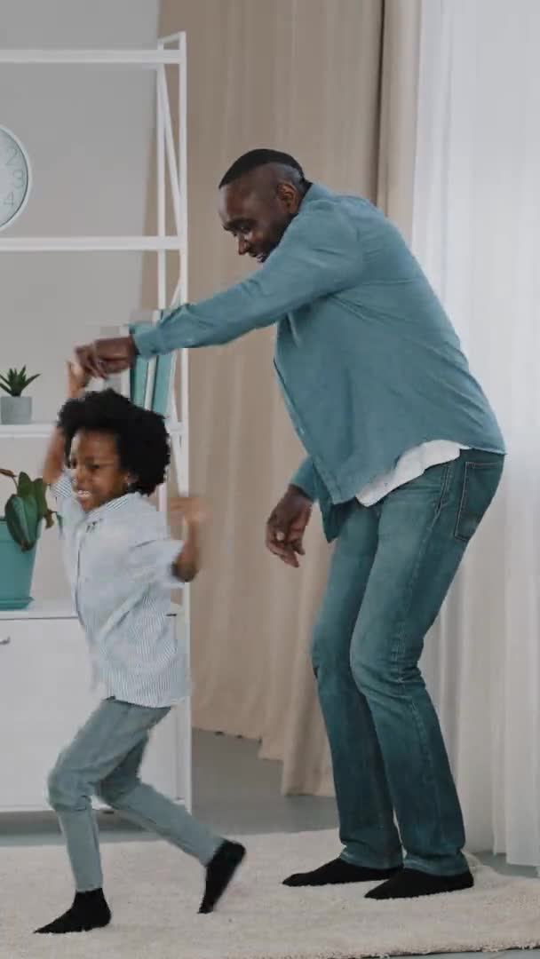 Vertical tiro maduro africano americano padre divertirse con la pequeña hija bailando a la música dinámica mover activamente jugar fines de semana en casa divertido niña saltando aferrándose a papá feliz familia — Vídeo de stock