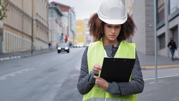 Ingeniera afroamericana en chaleco de seguridad casco de ingeniería hardhat de pie en la ciudad comprobando el tráfico por carretera señalando con el dedo en la distancia comprueba los datos en el portapapeles. Mujer profesional logística — Vídeo de stock