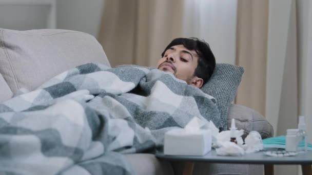 Индийский бородатый грустный измученный мужчина лежит на домашнем диване плохо покрытое одеяло страдает от лихорадки симптомов коронавируса концепции глядя на лекарства на столе устал от лечения covid19 сезонного гриппа — стоковое видео