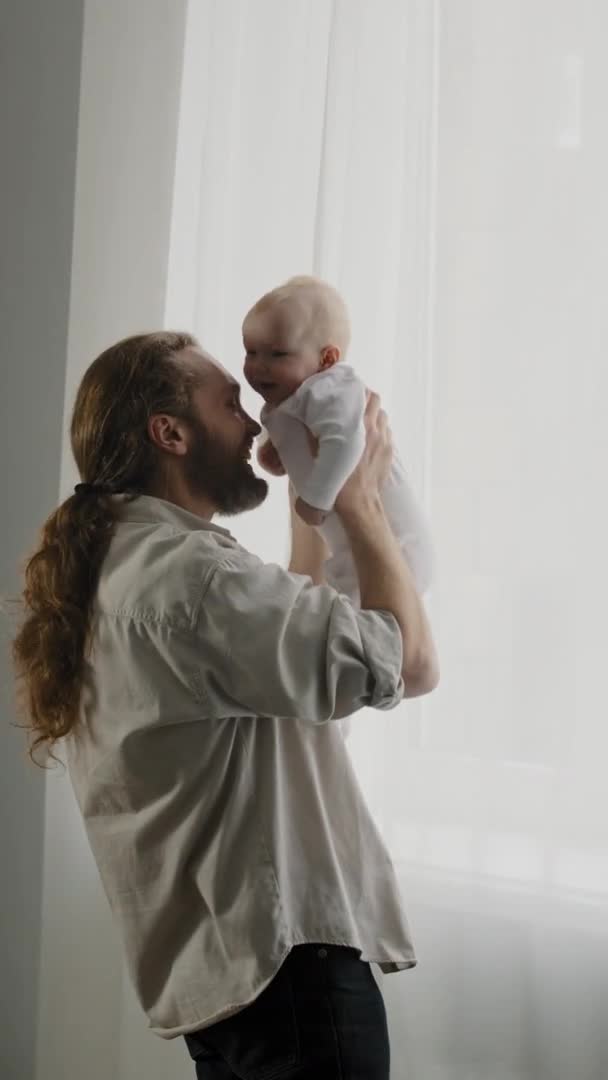 Vue verticale caucasien barbu père tenant bébé nouveau-né à la maison près de rideaux de fenêtre papa toucher le nez avec bébé chatouiller enfant enfant jouer avoir du plaisir jette fille soins papa avec fils collage — Video