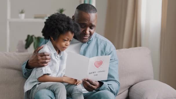 Volwassen afrikaanse amerikaanse man met schattig kind meisje knuffelen zitten in kamer op bank vader leest wenskaart liefhebbende dochter gemaakt geschenk met eigen handen voor verjaardag of vaders dag vader gelukkig knuffelen baby — Stockvideo