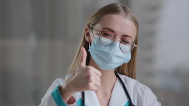 Caucasian woman doctor in glasses and face medical mask in clinic hospital looking at camera showing thumb up recommends vaccination health insurance immunization success end pandemic like gesture — Stock Video