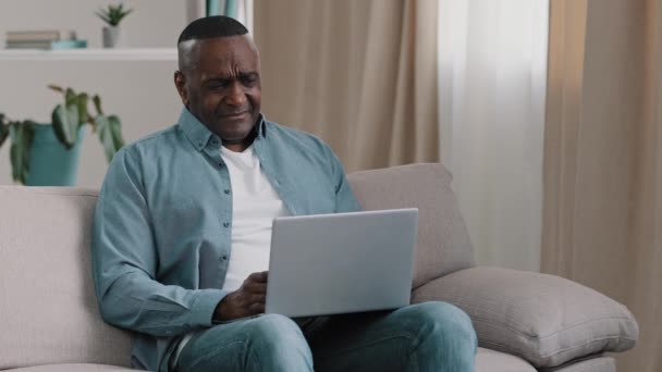 Volwassen gefrustreerde Afrikaanse Amerikaanse man zit op de bank in de kamer kijkt naar laptop scherm lezen slecht nieuws op e-mail gevoel gefrustreerd bezorgd over fout probleem faillissement mislukte resultaat ontslag — Stockvideo