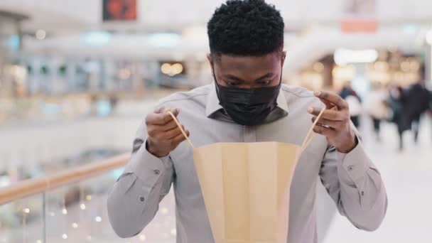 Κοντινό πλάνο νεαρός Αφροαμερικάνος άνδρας σε προστατευτική ιατρική μάσκα στέκεται στο εμπορικό κέντρο αρσενικό πελάτη αγοραστή γενέθλια άνθρωπος ψάχνει σε πακέτο με νέο δώρο αγοράς έκπληκτος συναίσθημα χαρά σοκ ευτυχία — Αρχείο Βίντεο