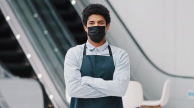 Afro-Amerikalı adam restoranda çalışan, maske takan, hastane maskesinde çalışan, kamera görüntüsü veren, iş yerinde önlük giymiş garson tezgâhtarı, salgın hastalık salgını olan karantinada dikiliyor. Küçük işletme