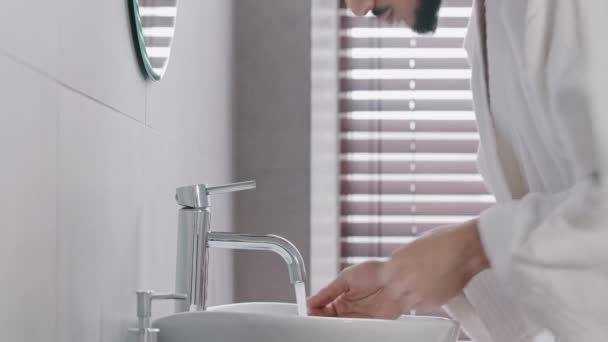 Vista lateral árabe español indio árabe hombre abre grifo cerca de lavabo en casa en el baño del hotel lava la cara con agua fría caliente belleza ritual masculino higiene chico en albornoz lavado facial húmedo — Vídeos de Stock