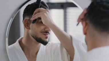 Arap İspanyol bir adam aynaya bakıyor saç fırçasıyla saçını tarıyor banyoda hazırlanıyor banyo aynası yansıması saç kesiminde güzellik rutini randevuya hazırlanıyor