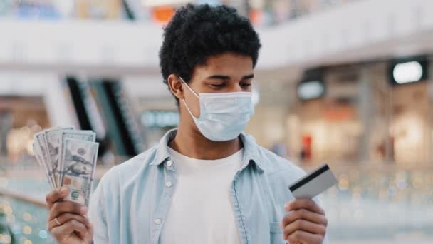 African american man guy male winner wears medical face mask holds bank card and dollars money in hands makes choice between cash and electronic finance rejoices in cashing out looking at camera — Stock Video