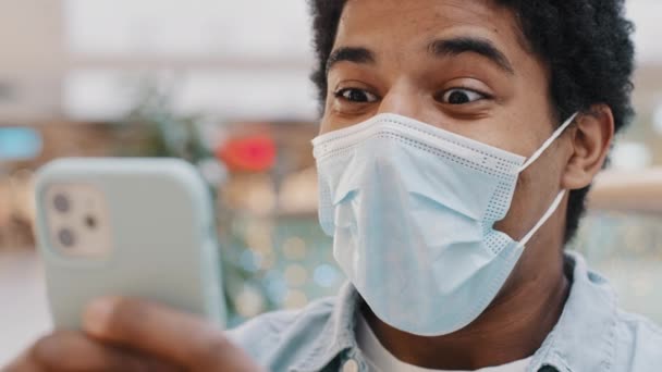 Hombre enmascarado afroamericano en máscara médica mensaje sorprendido recibido gana apuestas en línea mirando el teléfono móvil con los ojos abiertos sorpresa final pandemia noticias inesperado buen resultado en el teléfono inteligente — Vídeo de stock