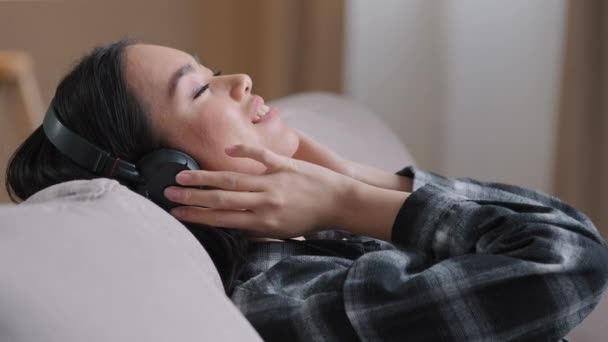 Nyugodt ázsiai koreai millenniumi nő fülhallgató ül kényelmes otthoni kanapén csukott szemmel hallgat kedvenc zene audio dal hang ritmikus kellemes kompozíció élvezze fülhallgató — Stock videók